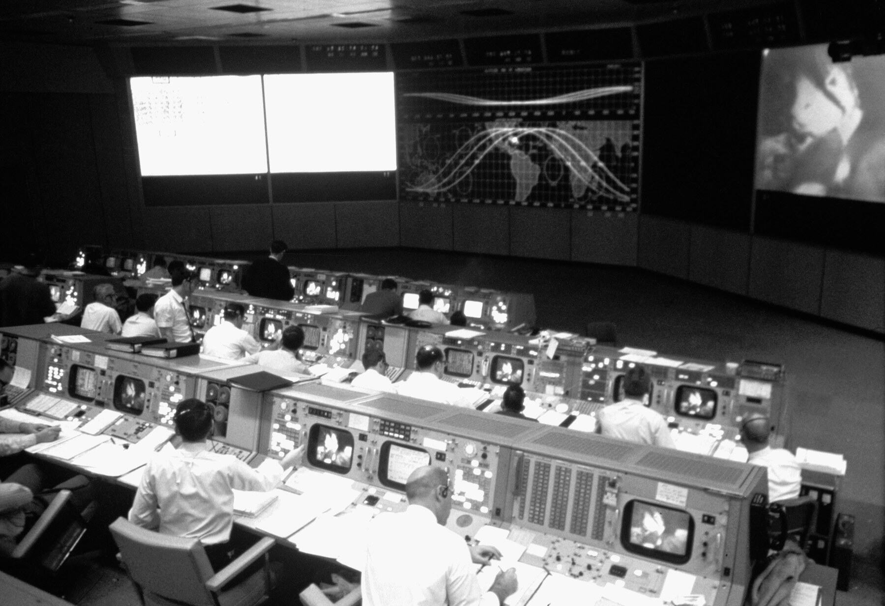 Salle de contrôle de mission Apollo : rangées de consoles face au grand tableau lumineux.
